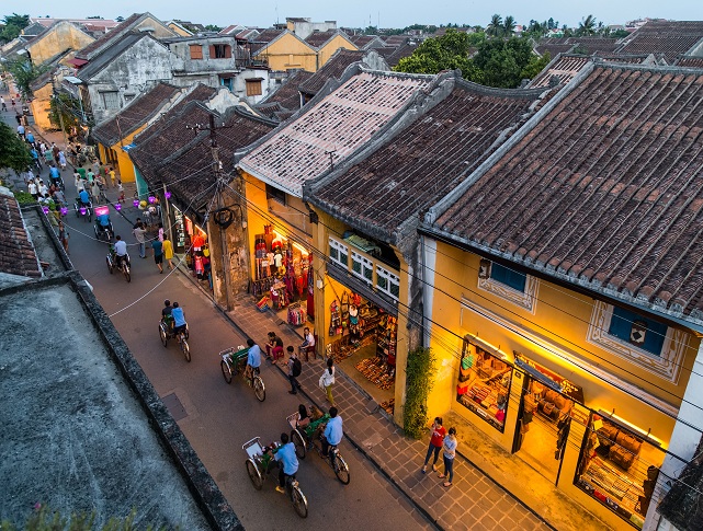 Aerial view of Hoi An Ancient Town with yellow traditional houses, lantern-lit streets, bicycles, and people walking through the historic old quarter at dusk.