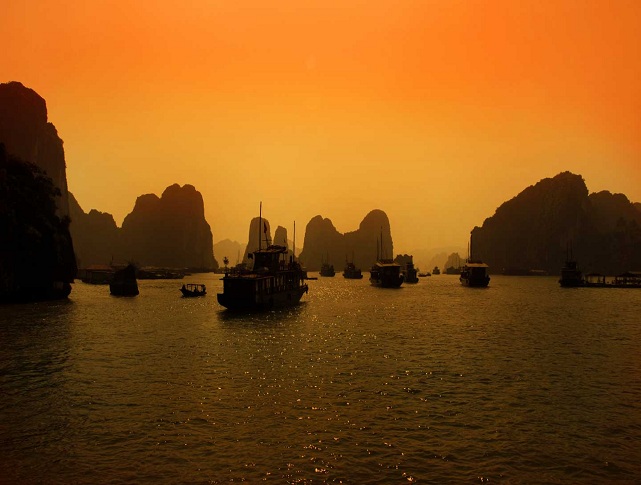 Boats sailing across Ha Long Bay during a golden sunset surrounded by limestone islands.
