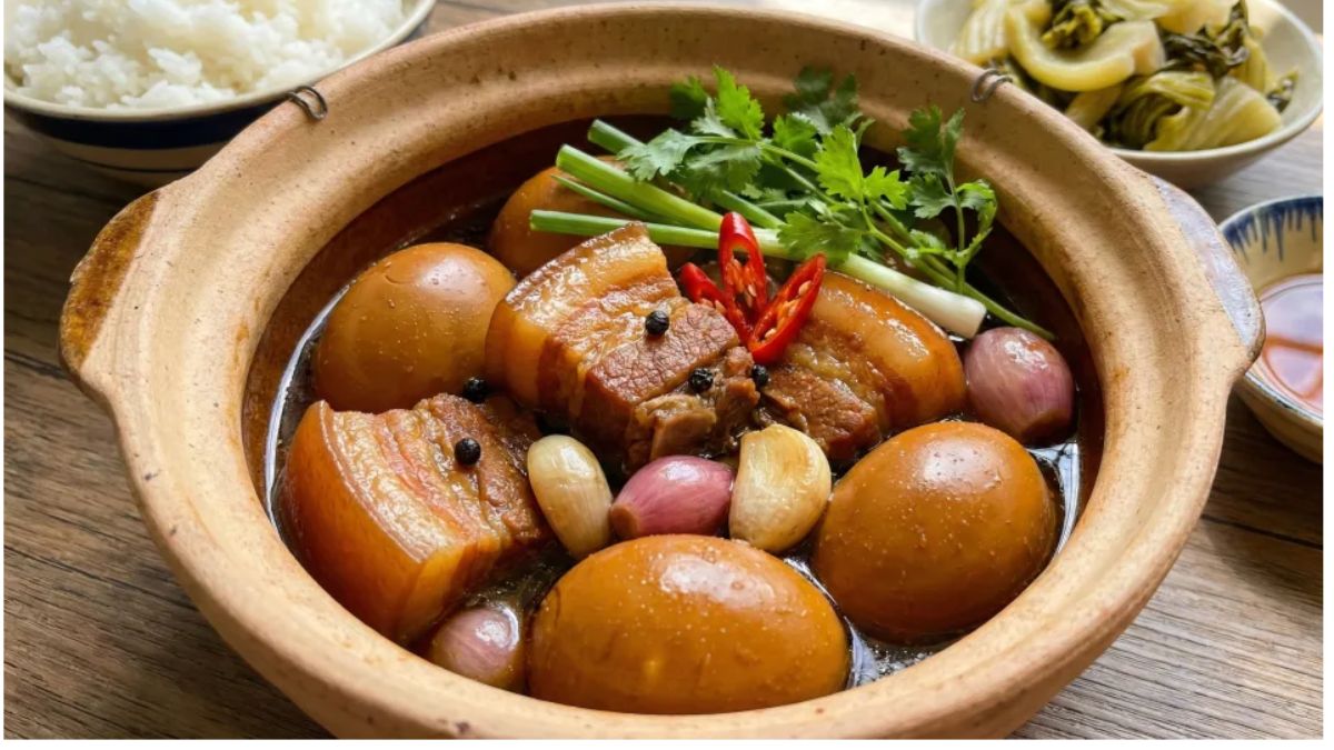 A traditional Vietnamese clay pot of braised pork belly with eggs (Thịt Kho Tàu), garnished with fresh herbs, chili, and shallots, served with steamed rice and pickled vegetables.
