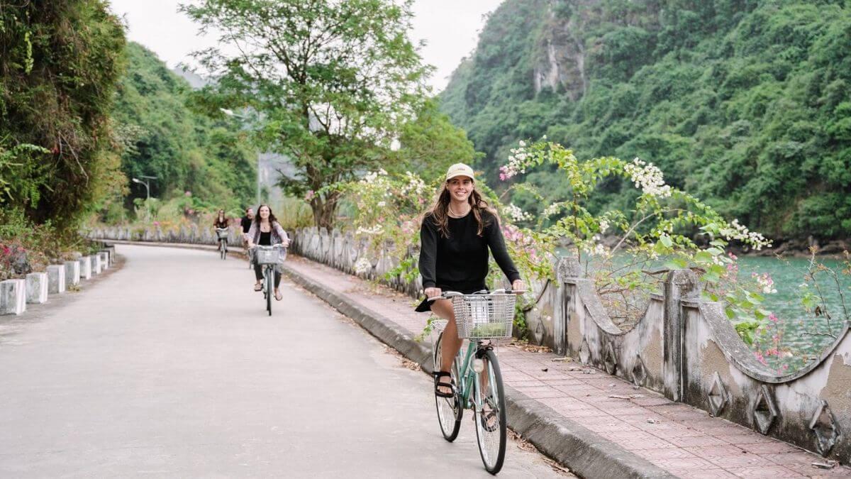 A smiling person rides a bicycle with a front basket along a quiet road beside a turquoise river, while two other cyclists follow behind. The road is lined with flowering vines, trees, and lush green limestone hills.
