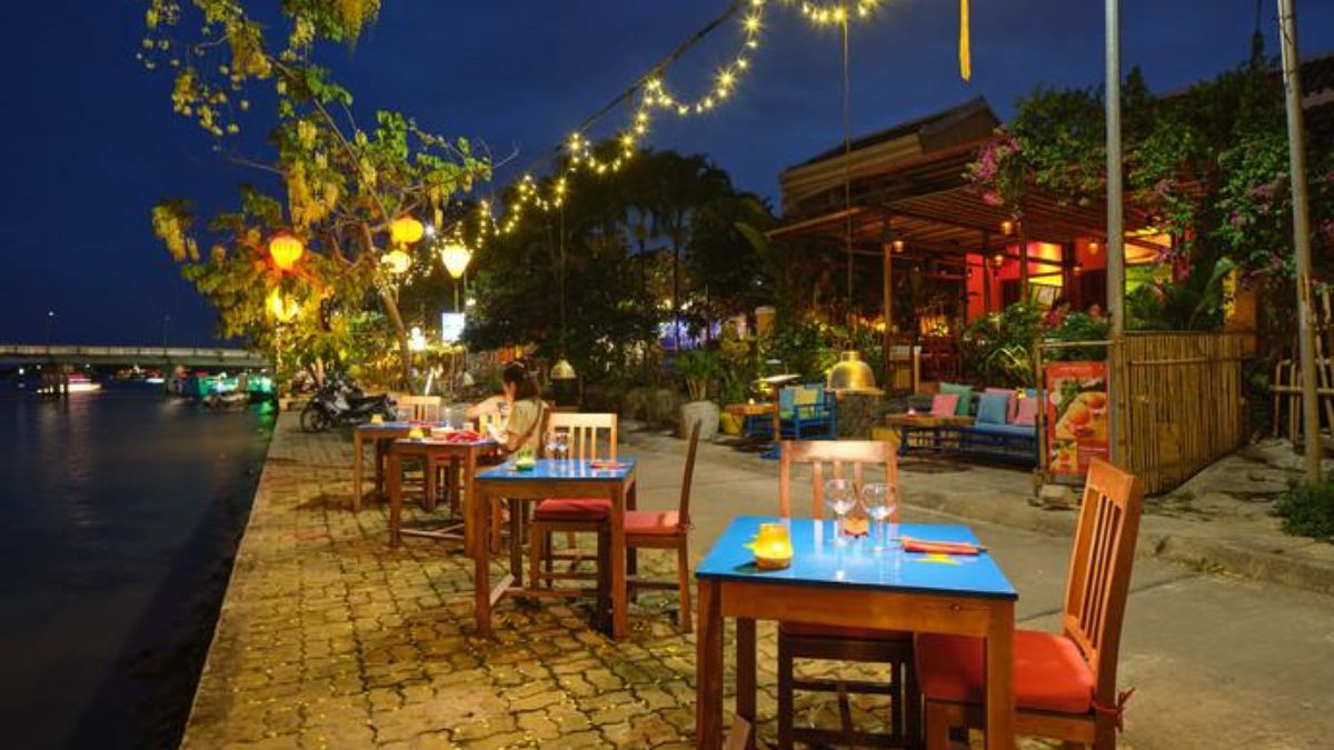 An outdoor riverside restaurant along the Hoai River in Hoi An, featuring candlelit tables, string lights, colorful lanterns, and a romantic evening atmosphere beside the water.