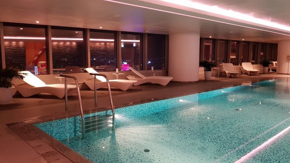 A modern indoor swimming pool at a luxury hotel in Hanoi, featuring crystal-clear water, comfortable lounge chairs, and large floor-to-ceiling windows with city views.