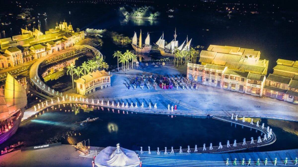 An aerial night view of the Hoi An Memories Show, featuring a spectacular outdoor stage with illuminated traditional architecture, boats, performers in colorful costumes, and a scenic waterfront setting.
