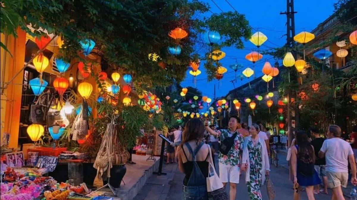 Visitors walk through the vibrant Hoi An Night Market under colorful lanterns, browsing souvenir stalls and enjoying the lively evening atmosphere in the ancient town.