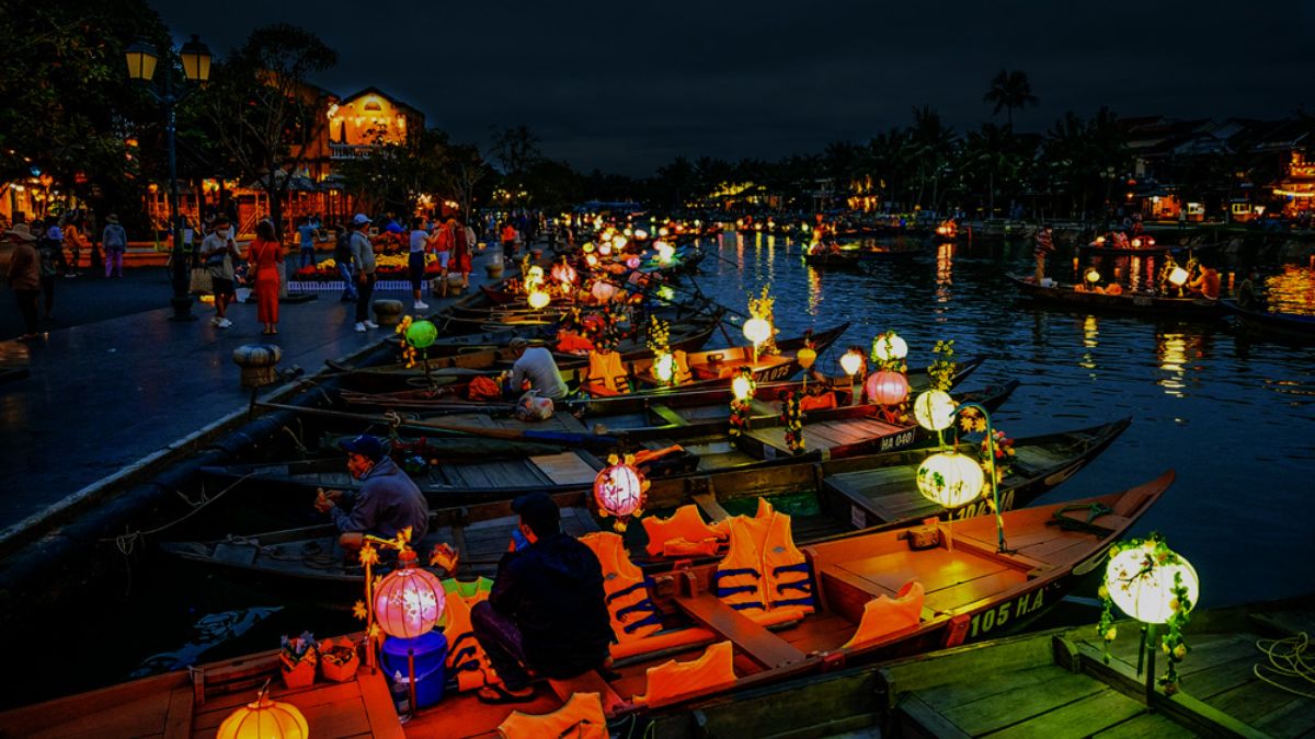 An outdoor riverside restaurant along the Hoai River in Hoi An, featuring candlelit tables, string lights, colorful lanterns, and a romantic evening atmosphere beside the water.
