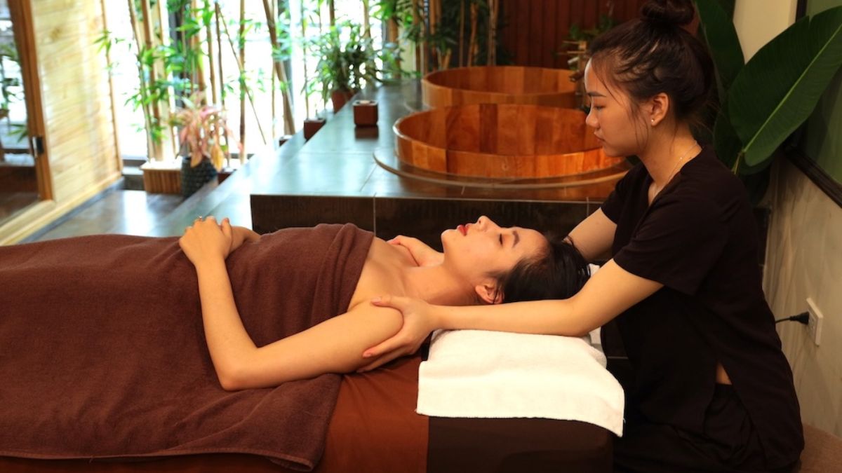 A guest receiving a relaxing neck and shoulder massage from a professional therapist at a tranquil spa in Hanoi, with wooden soaking tubs and lush greenery in the background.