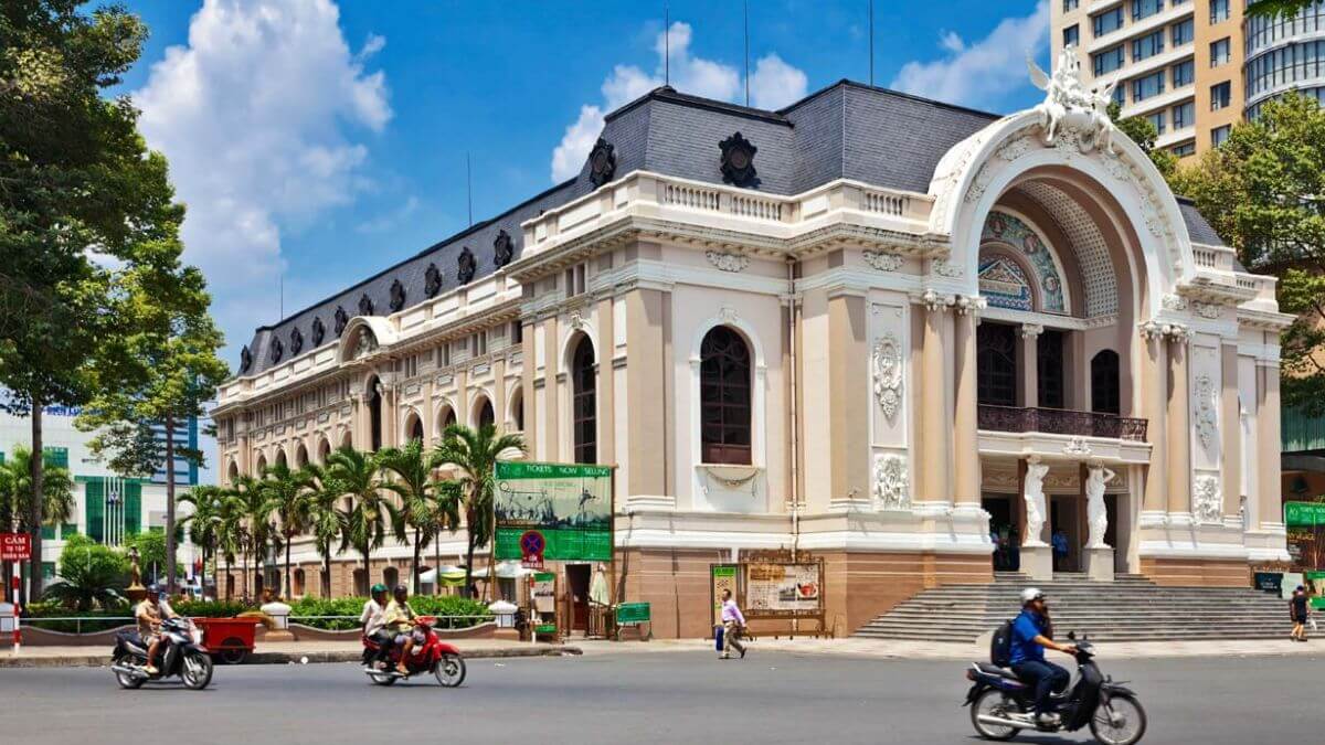 The Saigon Opera House, a beautifully preserved French colonial landmark, stands along a busy boulevard in downtown Ho Chi Minh City as motorbikes pass by.