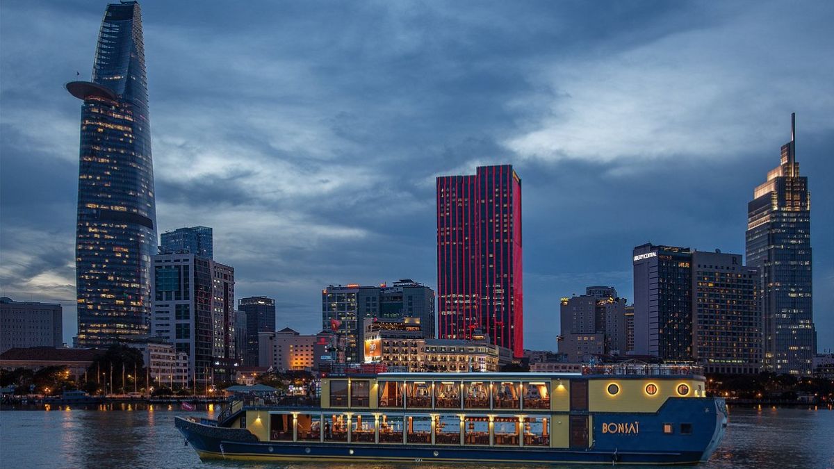A Saigon River cruise sails through Ho Chi Minh City at night, surrounded by illuminated skyscrapers, colorful city lights, and reflections shimmering on the water.