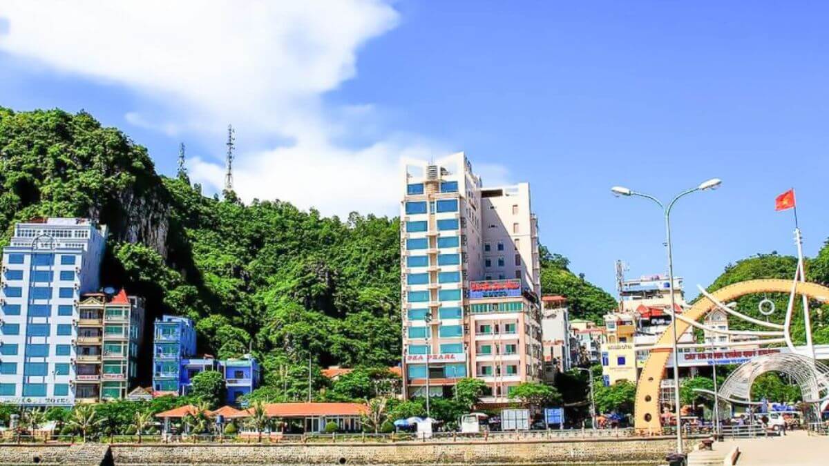 A waterfront view of Cat Ba Town features modern hotels, lush limestone hills, and the entrance to the Cat Ba tourist pier under a bright blue sky.