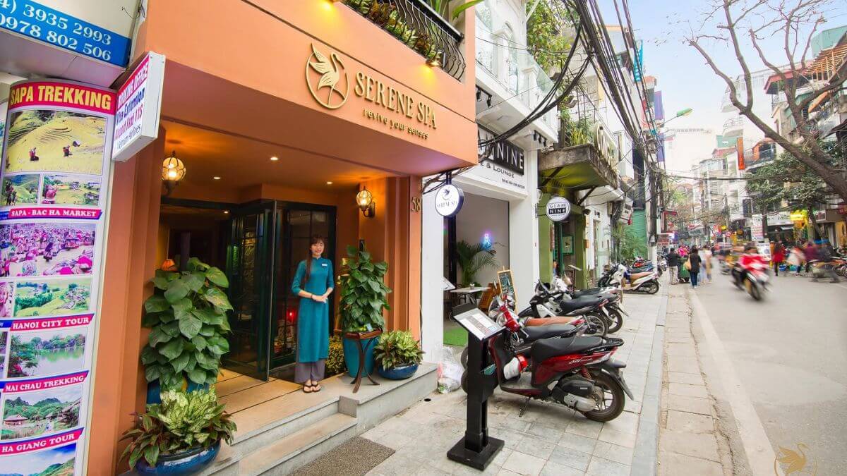 The entrance of Serene Spa in Hanoi's Old Quarter, featuring warm-toned architecture, lush potted plants, and a staff member welcoming guests at the door.
