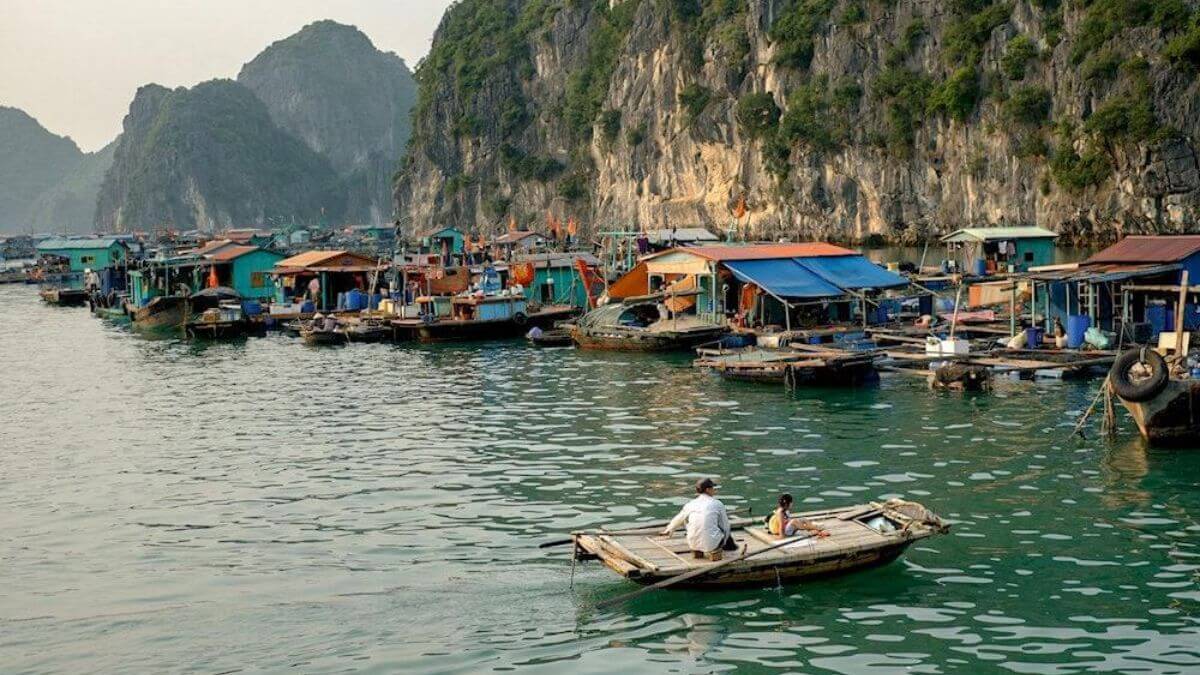 A traditional floating fishing village in Lan Ha Bay features colorful floating houses, calm emerald waters, and towering limestone cliffs, with a small wooden boat gliding across the bay.