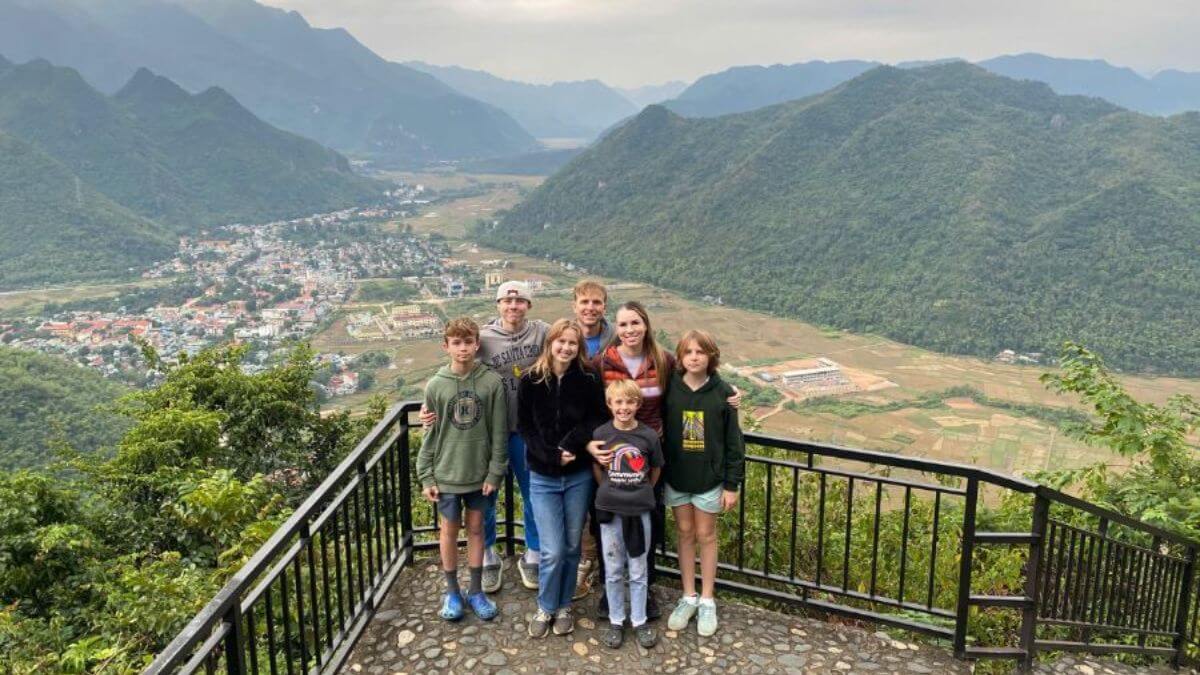 A family of international travelers poses at a scenic viewpoint in Cuc Phuong National Park, overlooking a lush valley surrounded by limestone mountains and rural landscapes.