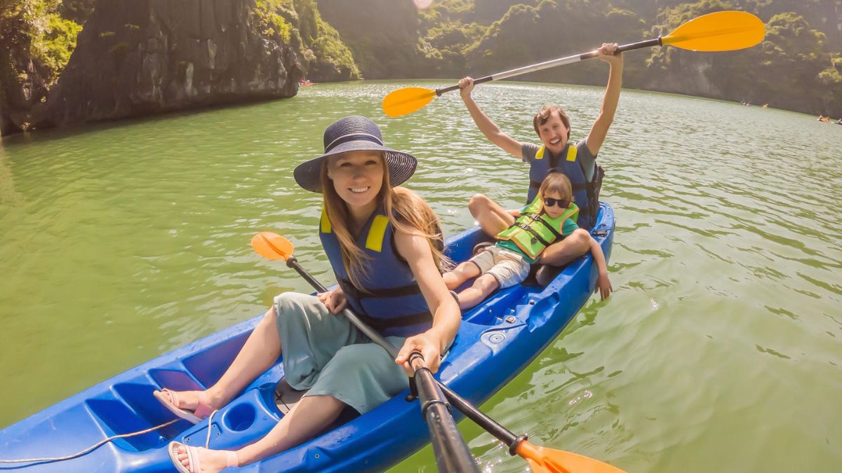Foreign family kayaking in Ha Long Bay, Vietnam, with a young child sitting between parents as they paddle through emerald waters surrounded by limestone cliffs.