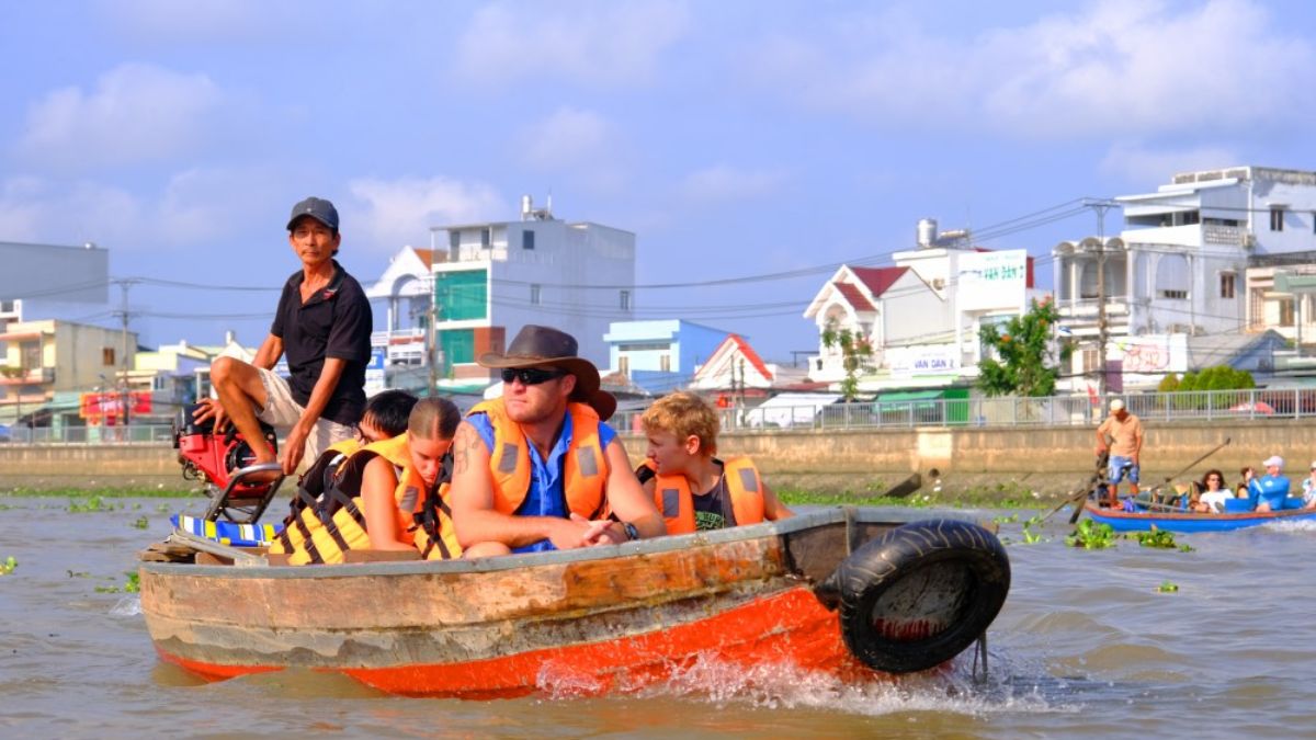 Western family with children wearing life jackets rides a traditional boat through a floating market in Vietnam’s Mekong Delta, accompanied by a local boat driver.