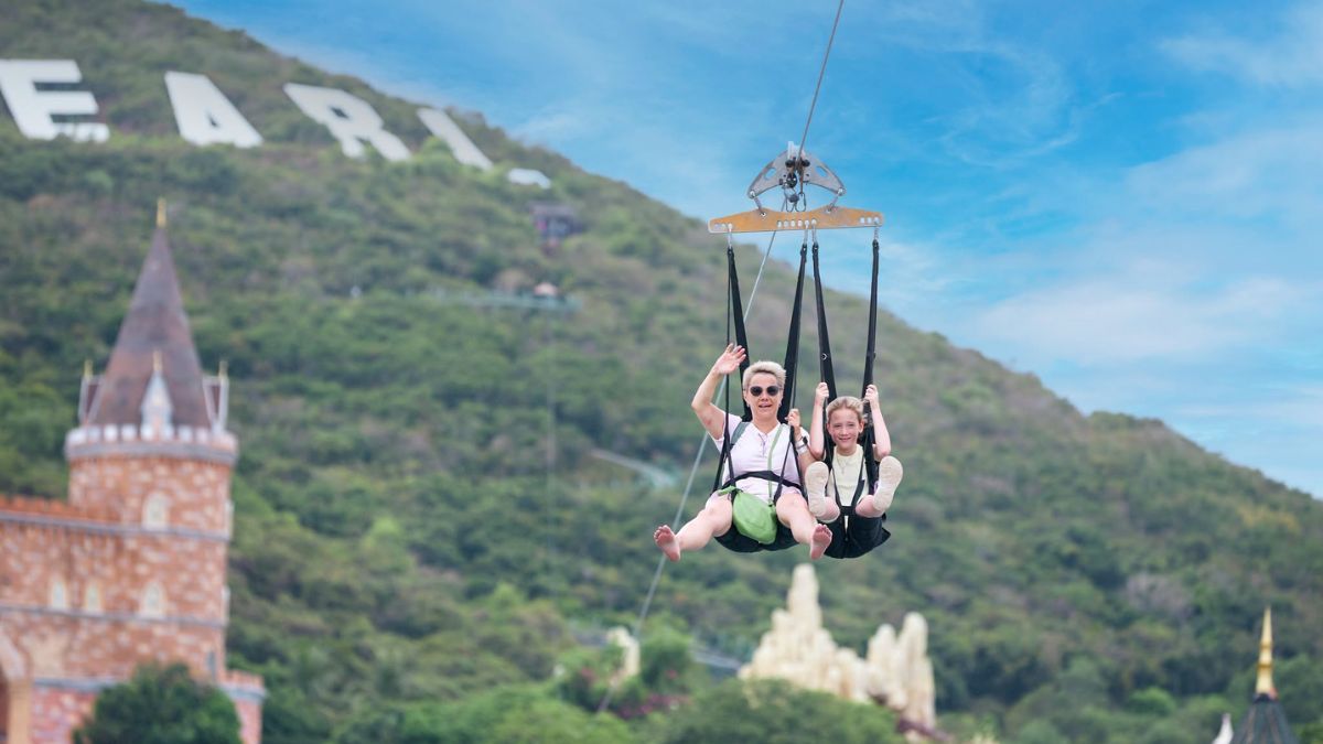 A smiling adult and a young girl ride a zipline attraction at VinWonders Nha Trang, soaring above the park with green mountains and castle-like architecture in the background.