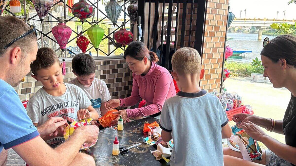 An international family and their children participate in a traditional lantern-making workshop in Hoi An, learning local craftsmanship under the guidance of a Vietnamese artisan.