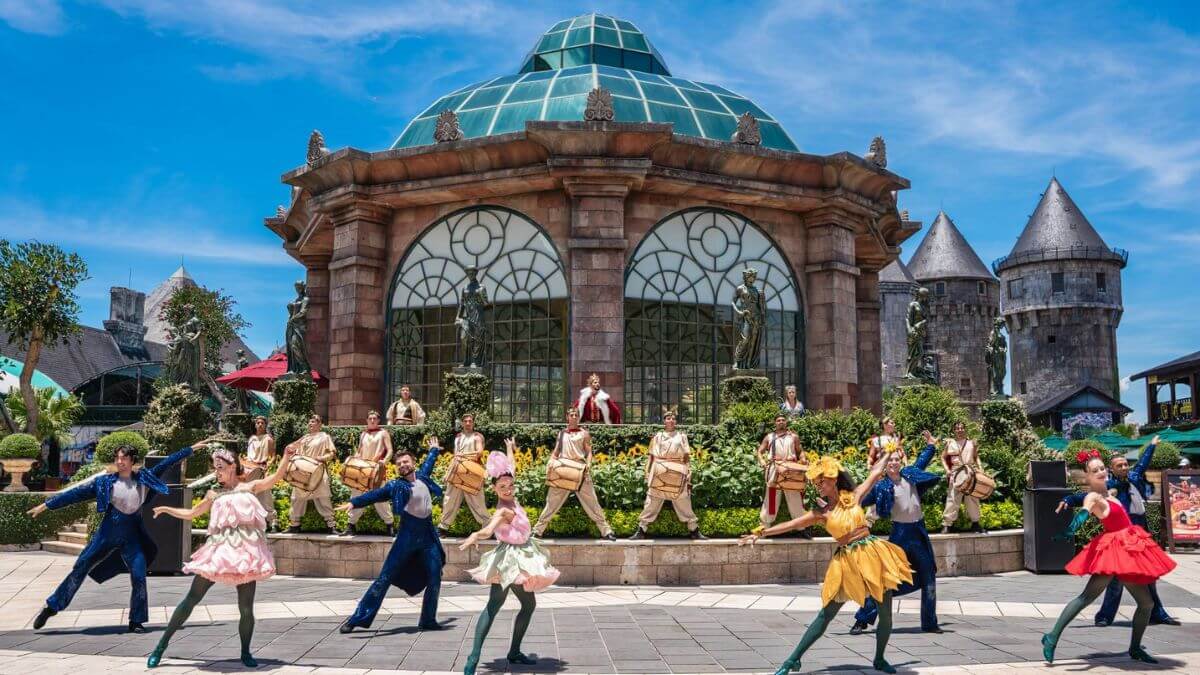 Performers in colorful costumes dance in front of a grand European-style building at Sun World Ba Na Hills, creating a lively atmosphere in the heart of the theme park