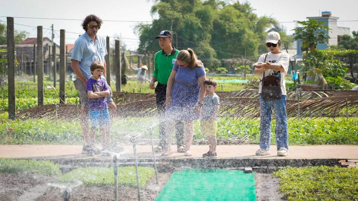 An international family visits Tra Que Vegetable Village in Hoi An, watching a local guide demonstrate traditional irrigation techniques in a lush vegetable garden.
