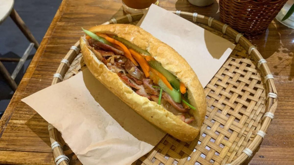 A close-up of a Vietnamese banh mi sandwich filled with grilled pork, pickled carrots, cucumber slices, and fresh herbs, served in a crispy baguette on a woven bamboo tray.