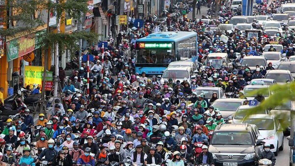 Heavy motorbike traffic at a busy intersection in Vietnam, with hundreds of riders waiting at a traffic light alongside cars and taxis during rush hour