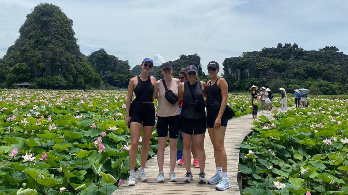 A group of four women in athletic wear stand smiling on a wooden boardwalk that weaves through a massive field of blooming pink lotus flowers, with limestone mountains in Ninh Binh in the background.