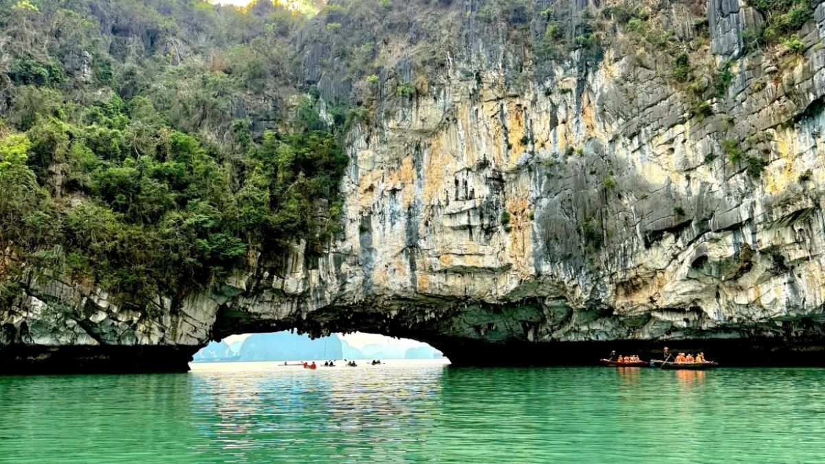 Bai Tu Long Bay featuring a dramatic limestone arch cave over emerald waters, with small boats exploring one of Vietnam’s most pristine natural landscapes.