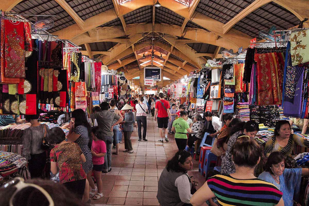 Inside Ben Thanh Market in Ho Chi Minh City with crowded aisles, colorful fabric stalls, souvenir shops, and visitors exploring the bustling indoor market.