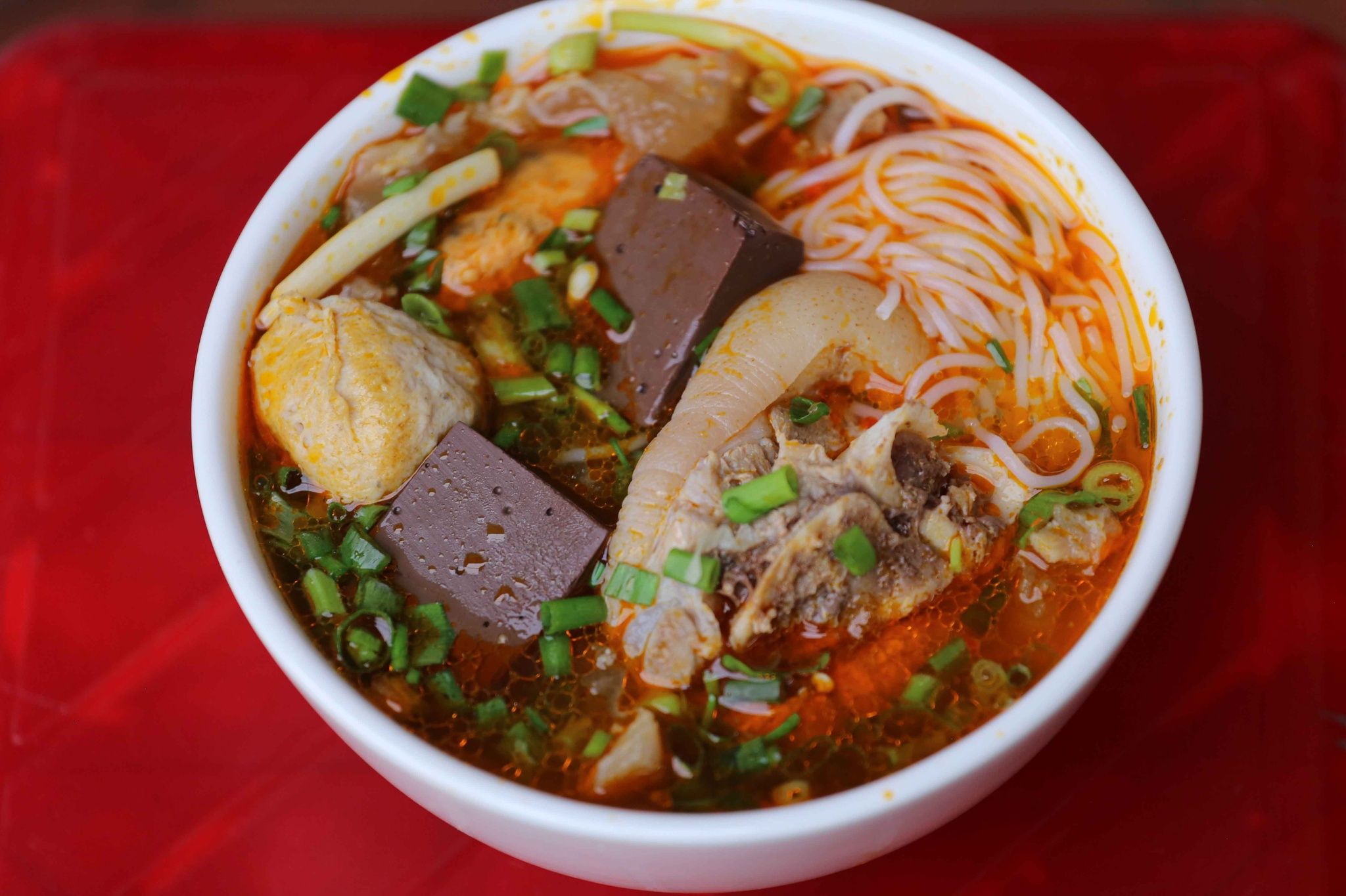 A bowl of Bun Bo Hue with spicy broth, rice noodles, beef, pork, blood cubes, and fresh herbs.