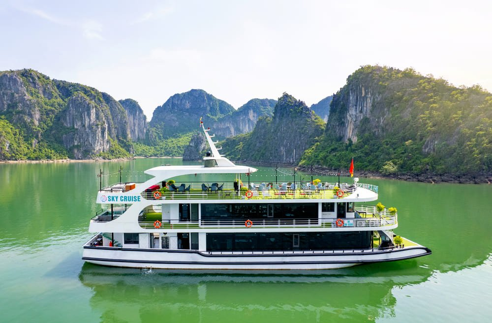 A modern white cruise ship sailing on the calm green waters of Ha Long Bay, surrounded by towering limestone mountains and lush greenery under a clear sky.