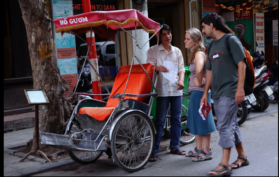 A cyclo driver speaking with two tourists beside a traditional cyclo on a street in Hanoi, Vietnam