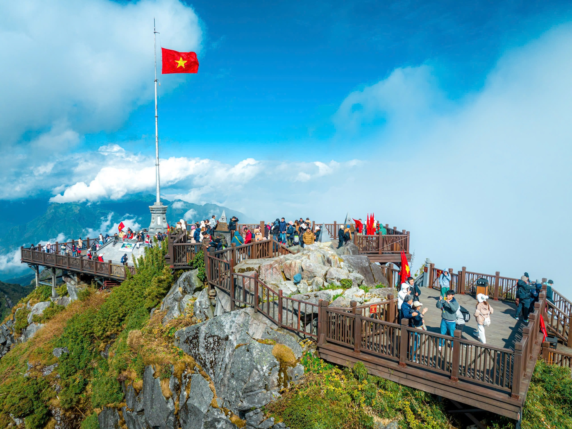 Visitors standing at the summit of Fansipan Mountain in Sapa with the Vietnamese flag waving above the clouds.