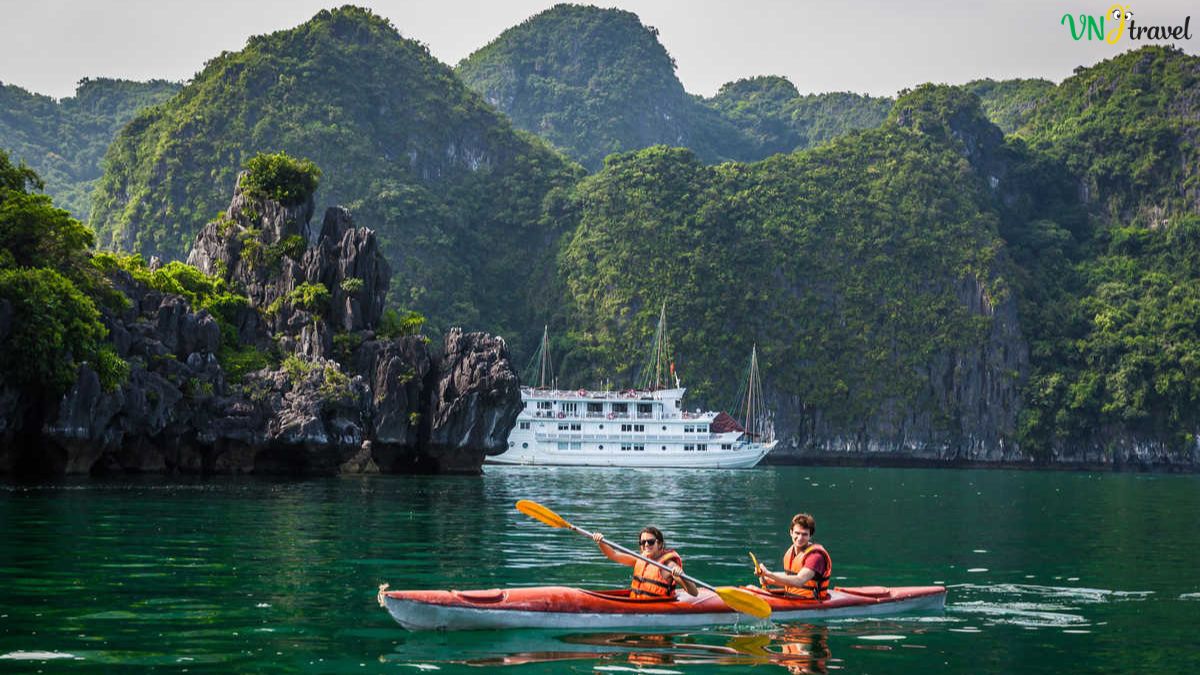 Tourists kayaking through the emerald waters of Ha Long Bay surrounded by limestone karst islands and a cruise ship in the background.