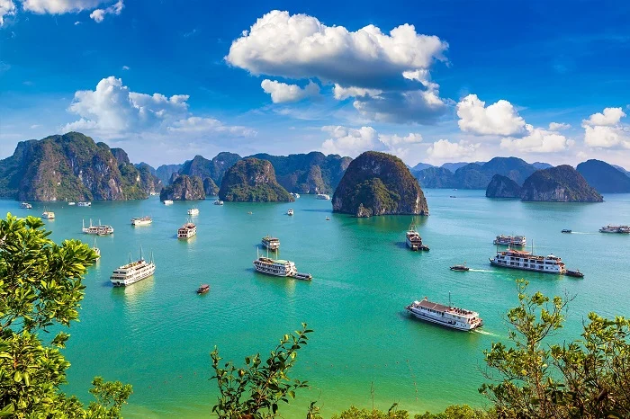 Boats cruise through turquoise waters surrounded by towering limestone islands under a bright blue sky with fluffy white clouds in Ha Long Bay, Vietnam.