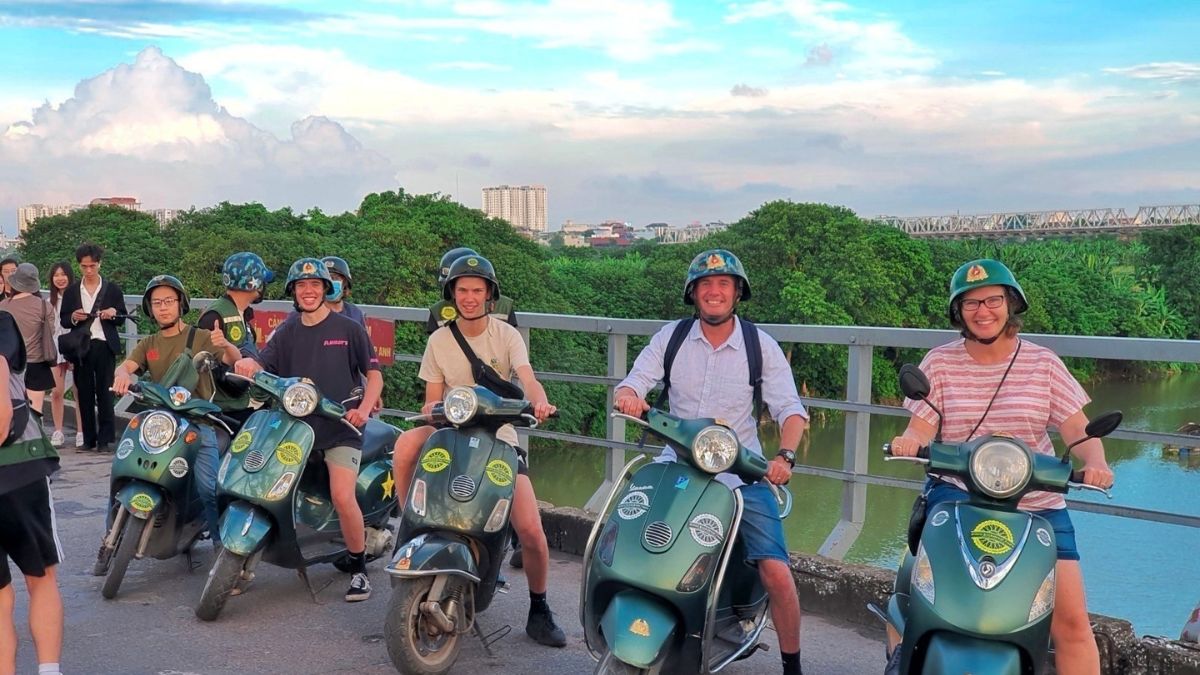 A group of tourists smiling while sitting on green Vespa scooters parked along a bridge railing over a river in Hanoi, wearing matching green helmets.