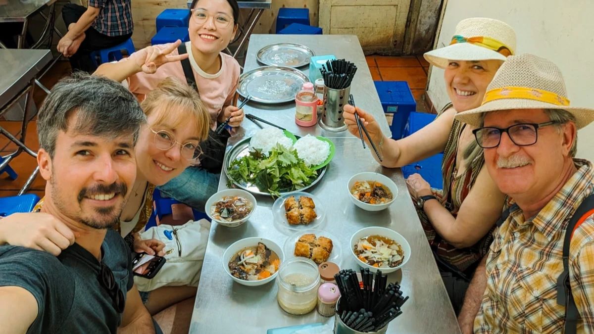 A high-angle selfie of a group of four tourists and their local guide smiling around a stainless steel table filled with bowls of Bun Cha, rice noodles, fresh herbs, and fried spring rolls.
