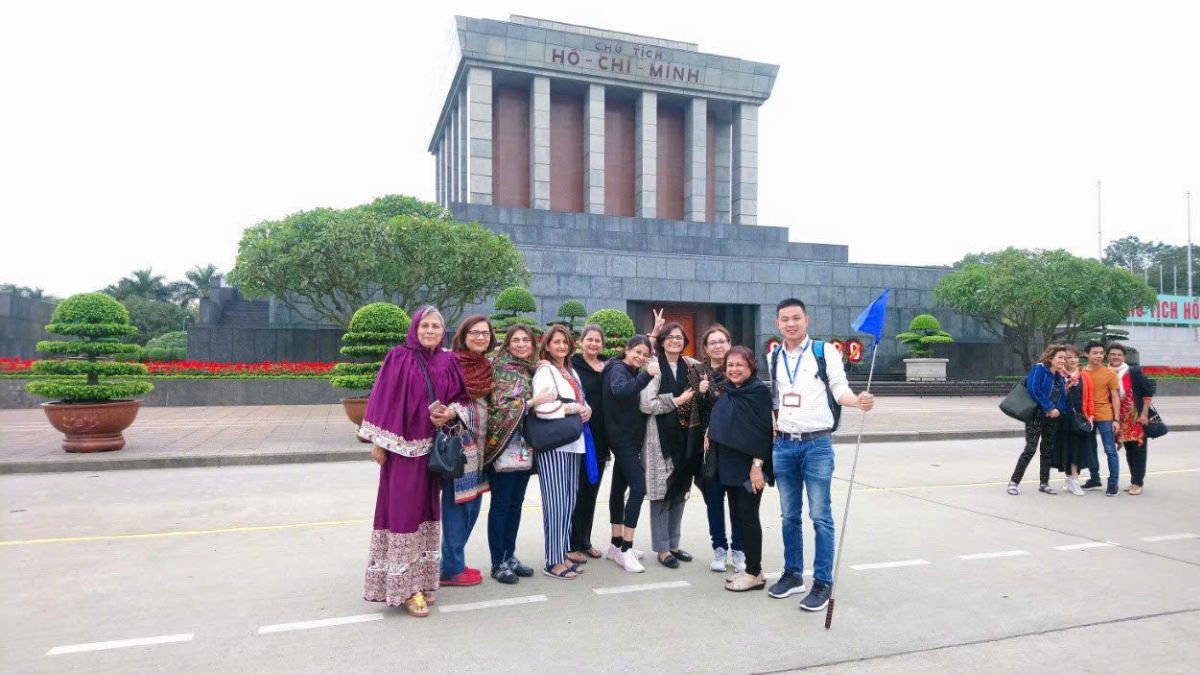A group of international tourists posing with their local guide in front of the Ho Chi Minh Mausoleum in Hanoi, Vietnam. The guide is holding a blue tour flag while the visitors smile for a souvenir photo.