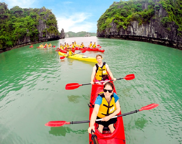 Tourists kayaking on emerald-green water in Ha Long Bay, surrounded by towering limestone cliffs covered in lush greenery, with several red and yellow kayaks lined up behind them.