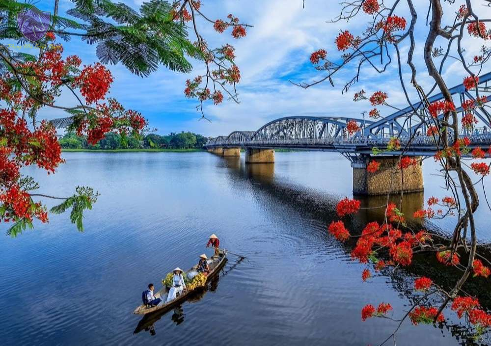 A peaceful view of the Perfume River in Hue with a traditional boat, blooming red flamboyant flowers, and Truong Tien Bridge in the background.