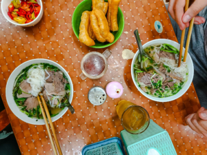 Two bowls of traditional Hanoi pho with beef, fresh herbs, and fried dough sticks on a wooden dining table.