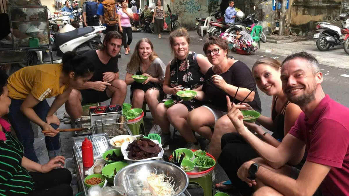 Tourists enjoying authentic Vietnamese street food at a small roadside stall in Hanoi, sitting on low plastic stools and sharing local dishes in a bustling urban alley.