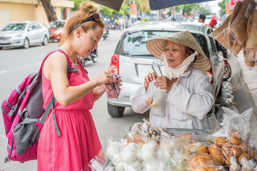 A tourist buying snacks from a street vendor wearing a traditional conical hat on a sidewalk in Vietnam