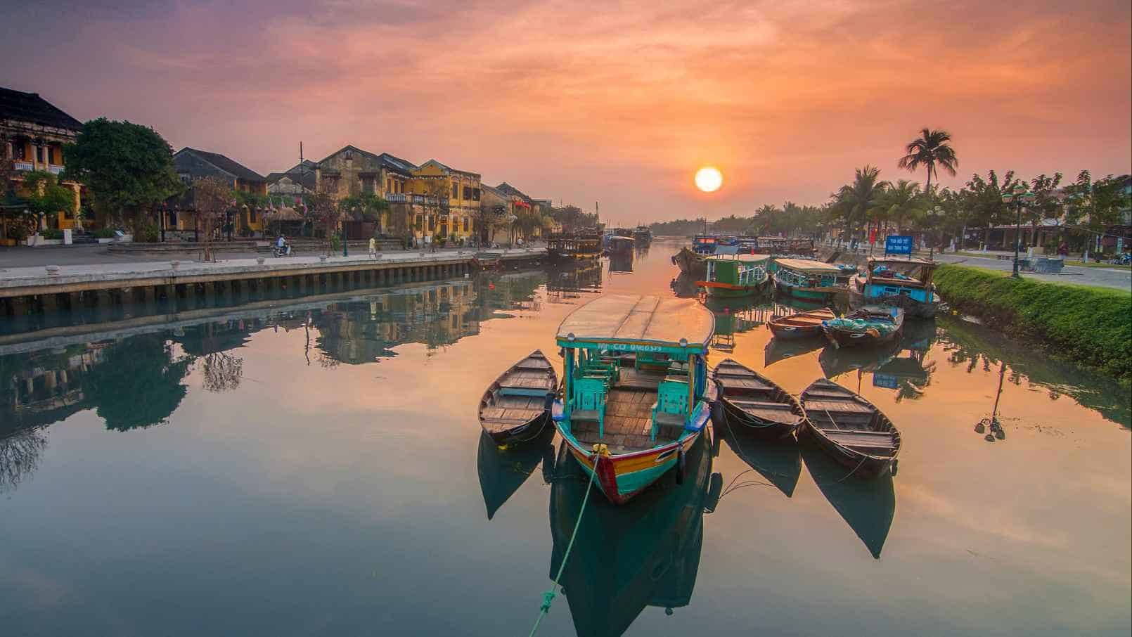 Boats resting along the Thu Bon River at sunset in Hoi An Ancient Town, with colorful reflections on the calm water and traditional riverside houses in the background.