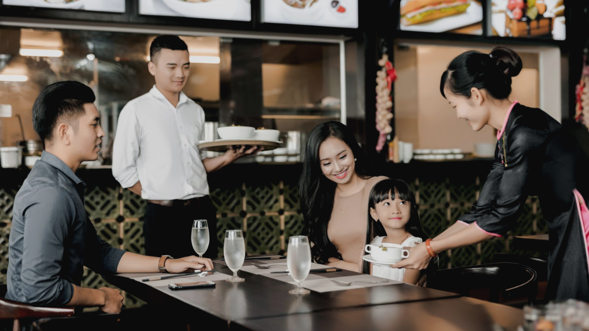A Vietnamese family dining at a modern restaurant while a waitress serves a bowl of soup to a young girl at the table.