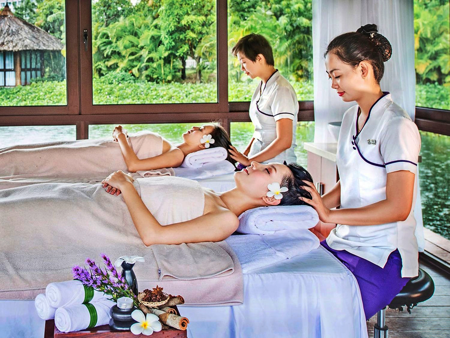Two women enjoying relaxing head massages from spa therapists at a luxury wellness resort surrounded by tropical greenery.