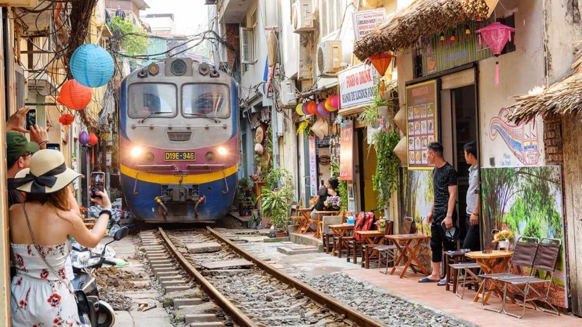 A narrow street in Hanoi's famous Train Street, where colorful cafés line the railway tracks. Visitors sit just inches from the rails, waiting as a train passes through the heart of the neighborhood, creating one of Vietnam's most iconic urban experiences.