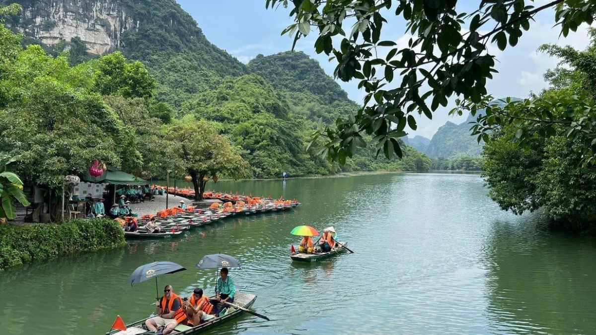 Small rowboats carrying tourists in orange life jackets cruise down a calm, emerald-green river surrounded by lush, forested limestone karst mountains at Trang An, Ninh Binh, Vietnam.