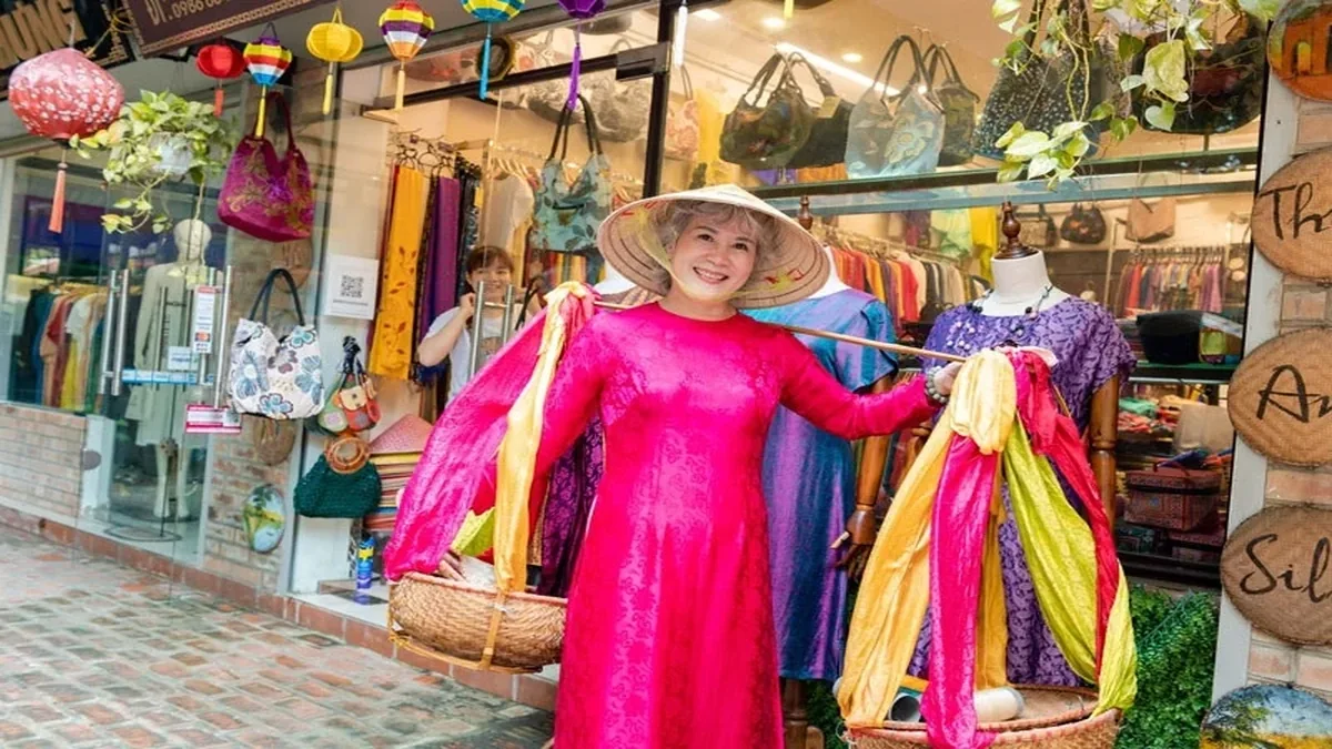 Woman wearing a traditional Vietnamese conical hat and bright pink áo dài displaying colorful silk fabrics at a silk shop in Van Phuc Silk Village, Hanoi, Vietnam.