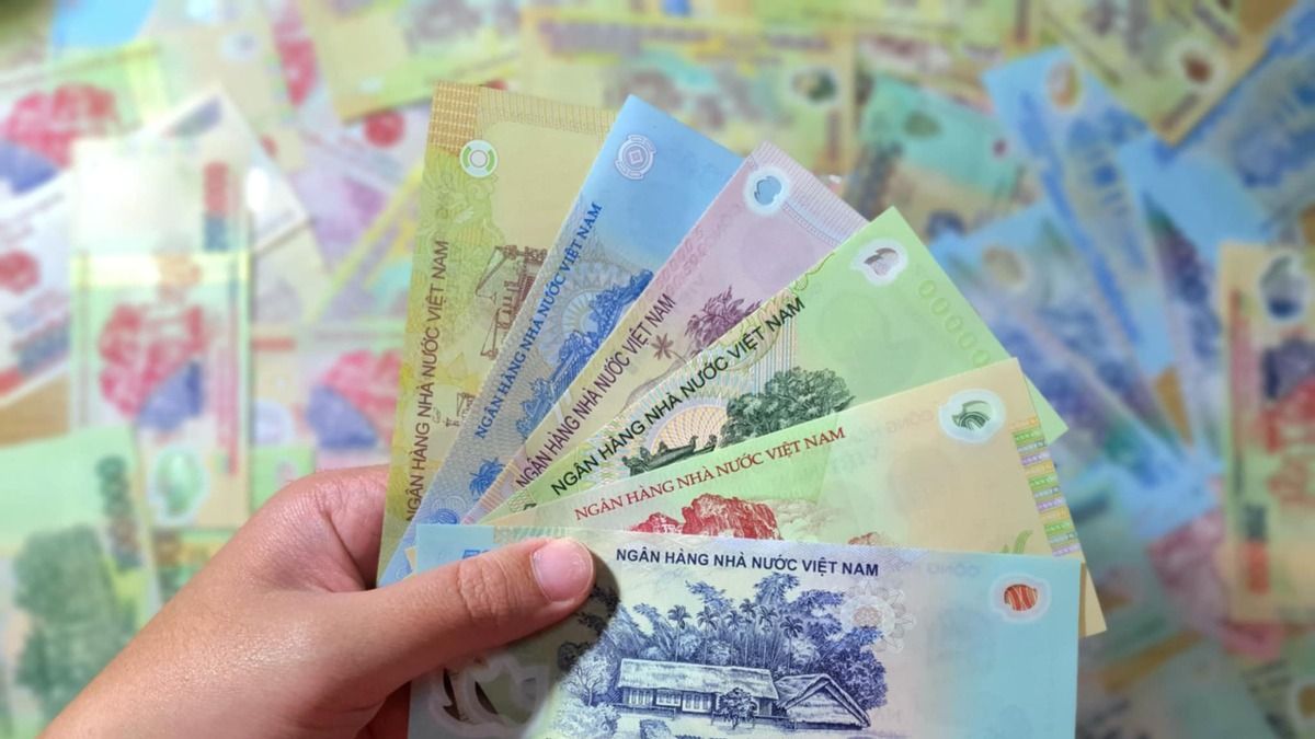 Hand holding colorful Vietnamese đồng banknotes with various denominations against a background of scattered Vietnam currency.