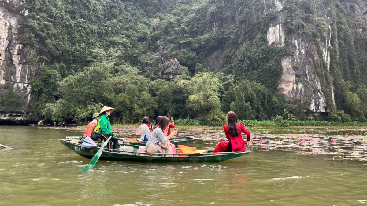 Visitors enjoy a traditional sampan boat ride through the scenic limestone mountains and lotus-covered river in Tam Coc, Ninh Binh, Vietnam.