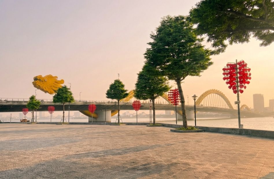 A wide paved waterfront promenade along the Han River in Da Nang, Vietnam, lined with green trees and red heart-shaped lantern posts,
